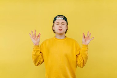 Kazaklı ve şapkalı genç adam sarı arka planda meditasyon yapıyor. Gözleri kapalı ve sakin bir yüzü var..