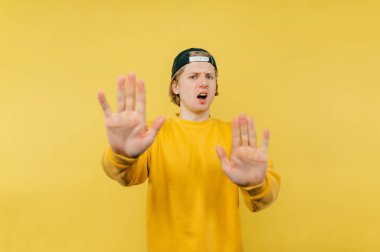 Tatmin olmamış genç bir adam kameraya bir jest yapıyor ve sarı arka planda şaşırmış bir yüzle bakıyor..
