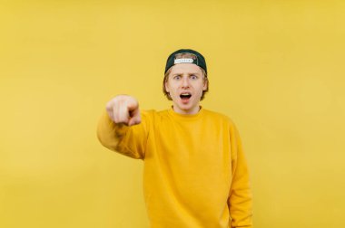 Şok olmuş, sarı tişörtlü ve şapkalı bir adam sarı bir arka planda, hoşnutsuz bir yüzle kamerayı işaret ediyor..