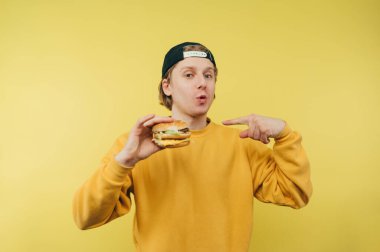 Sarı arka planda şapkalı, elinde hamburgerin parmağını gösteren olumlu yüzüyle kameraya bakan şaşırmış bir gencin portresi. Fast food.