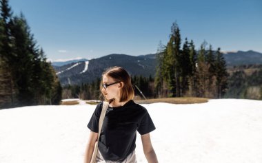 Tişörtlü güzel hippi kadın, kar manzaralı arka planda yürürken dağlarda duruyor ve ciddi bir yüz ifadesiyle başka yere bakıyor..