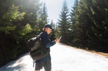 Yürüyüş sırasında sırtında bir sırt çantası ve elinde akıllı bir telefonla dağlarda kar altında dikilen sıradan giysili genç bir adam.