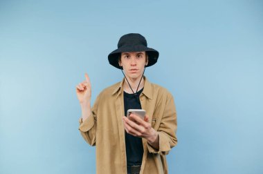 Panamalı konsantre hipster adam elinde bir akıllı telefon tutuyor ve mavi arka planda parmağını işaret ediyor..
