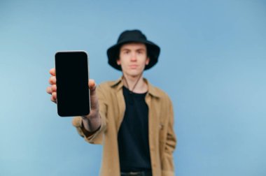 Ciddi bir genç adam, günlük giysiler içinde, gömlek ve panama giyiyor, mavi bir arka planda duruyor, ciddi bir yüzle kameraya bakıyor ve siyah ekranlı bir akıllı telefon ekranı gösteriyor.