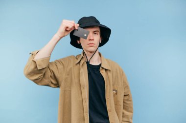 Yakışıklı genç hippi, elinde kredi kartıyla kameraya poz veriyor. Mavi arka planda ciddi bir yüzü var..