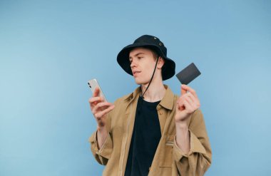 Panamalı ve elinde banka kartı olan tişörtlü bir adam mavi arka planda duruyor ve akıllı telefon kullanıyor.