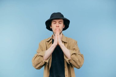 Panama ve gömlek giyen yakışıklı genç adam mavi arka planda duruyor ve gözleri kapalı, izole bir şekilde dua ediyor..