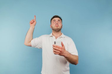Beyaz tişörtlü yetişkin bir adam elinde akıllı bir telefonla mavi arka planda duruyor ve parmağını boş bir yere doğrultuyor.