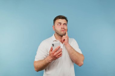 Elinde akıllı telefon olan düşünceli bir adam mavi bir arka planda duruyor ve başka tarafa bakıyor ve düşünüyor..