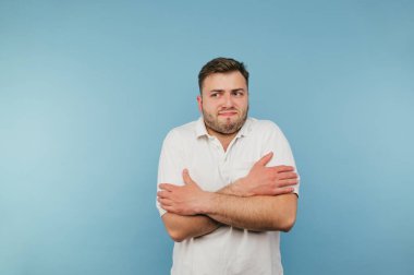 Beyaz tişörtlü donmuş adam mavi arka planda duruyor ve kendine sarılıyor ve hoşnutsuz bir yüzle başka tarafa bakıyor.