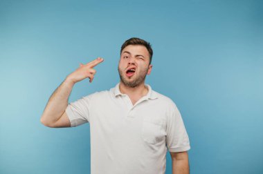 Beyaz tişörtlü duygusal yetişkin bir adam silahla parmağıyla kafasına ateş ediyor ve kameraya bakıyor, mavi arka planda bir intihar hareketi gösteriyor.