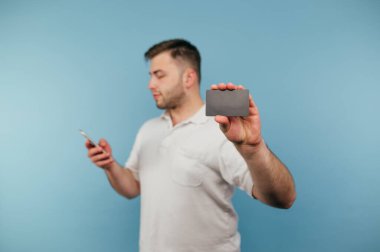 Beyaz tişörtlü adam akıllı telefon kullanıyor ve kameraya mavi arka planda siyah bir banka kartı gösteriyor..