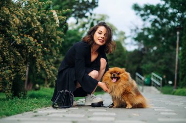 Çekici stil sahibi bir kadın parkta pomeranya köpeğiyle oturmuş yüzünde bir gülümsemeyle kameraya bakıyor..