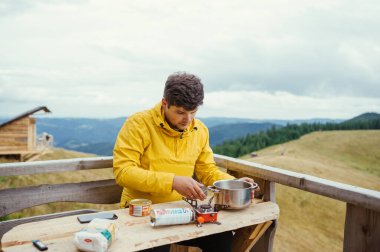 Bir erkek turist dağdaki bir kır evinin masasına oturur ve güzel bir manzaranın arka planına karşı ocakta yemek hazırlar..