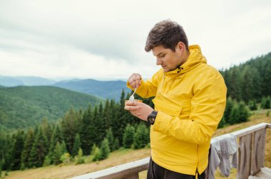 Erkek turist bir dağ kulübesinin terasında duruyor ve arka planda güzel manzaralı konserve yiyecekler yiyor..
