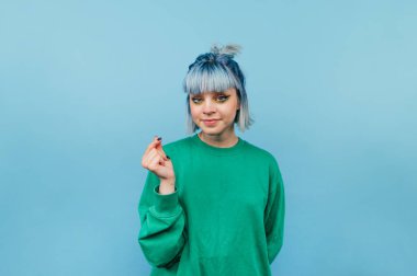 Renkli saçlı hoş bir bayan mavi arka planda duruyor ve yüzünde bir gülümsemeyle bir kalp hareketi gösteriyor..