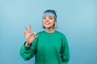 Renkli saçlı mutlu hippi kız mavi arka planda duruyor ve yüzünde bir gülümsemeyle iyi niyet gösteriyor..