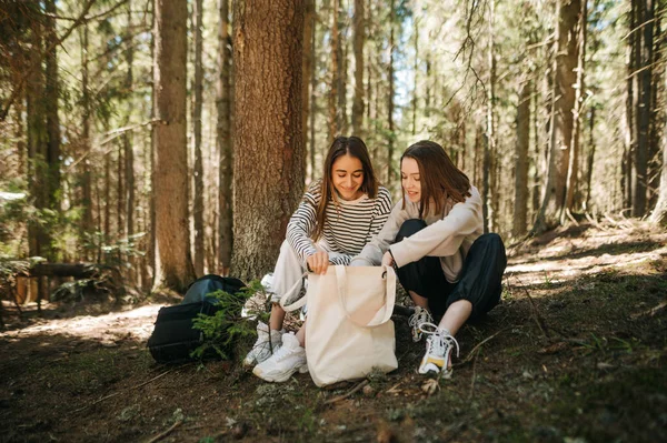 İki pozitif kız arkadaş ormanda oturuyor ve yüzlerinde bir gülümsemeyle ağaçların arkasındaki bir eko torbaya bakıyor..