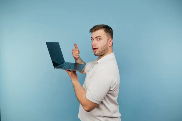 身穿白色T恤衫 手上拿着笔记本电脑 背景为蓝色的成年男子 摆出一副像照相机一样的姿势 看着摄像机 — 图库照片