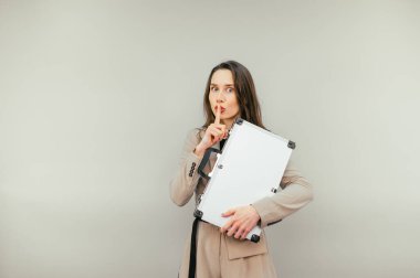 Serious business woman in a suit stands with a metal suitcase in his hands on a beige background and shows a gesture of silence.