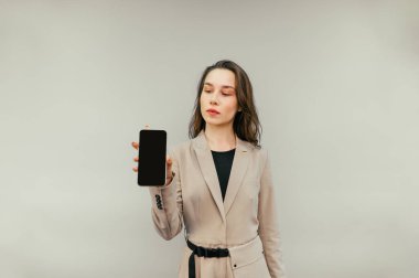 Ceketli güzel bir kadın bej bir arka planda duruyor ve siyah ekranlı bir akıllı telefon gösteriyor..