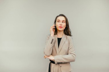 Ceketli hoş bir iş kadını yüzünde bir gülümsemeyle telefonda konuşuyor ve bej bir arka planda izole bir şekilde yüzünü çeviriyor..