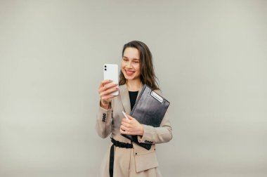 Ceketli güzel, pozitif bir kadın elinde tabletle bej bir arka planda duruyor ve yüzünde gülümseme olan bir akıllı telefon kullanıyor.