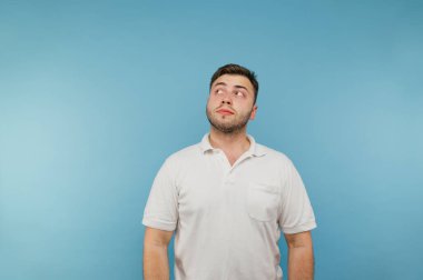 Beyaz tişörtlü ve tüylü dalgın bir adam mavi bir arka planda duruyor ve başka tarafa bakıyor ve düşünüyor..