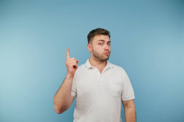 Smart man with bristles in a white T-shirt stands on a blue background and shows a finger up with a serious face.