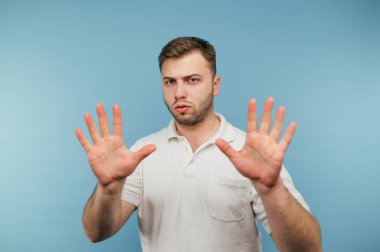 Beyaz tişörtlü ciddi bir adam mavi arka planda duruyor. Avuç içlerini ve kamerayı durdurup dikkatlice kameraya bakıyor.