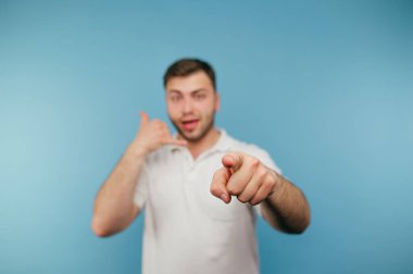 Beyaz tişörtlü olumlu adam bir çağrı işareti gösteriyor ve mavi arka planda parmağını kameraya gösteriyor..