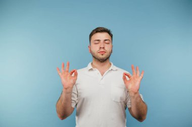 Beyaz tişörtlü yakışıklı yetişkin bir adam mavi arka planda duruyor ve gözleri kapalı meditasyon yapıyor.