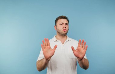 Beyaz tişörtlü bir adam mavi arka planda duruyor ve avuçlarını kameraya gösteriyor ve hoşnutsuz bir yüzle kameraya bakıyor..