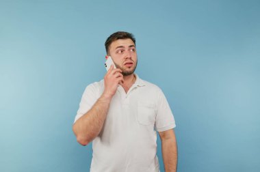 Beyaz tişörtlü yetişkin bir adam mavi arka planda duruyor, telefonla konuşuyor ve başka tarafa bakıyor..