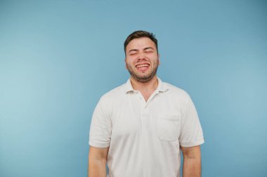 Beyaz tişörtlü neşeli adam mavi arka planda duruyor ve dişlerini göstererek gülüyor.