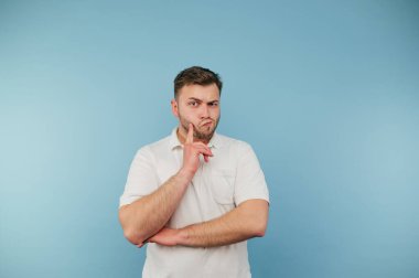 Bearded man in a white T-shirt stands on a blue background and looks at the camera with a pensive face.