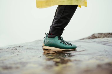 Dağlardaki su birikintisinde duran yeşil çizmeli bir erkek turistin bacakları beyaz arka planda bir kayanın üzerinde.