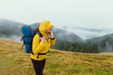A man hiker in a yellow jacket and with a backpack on his back stands in the mountains on a background of misty views and goes hiking.