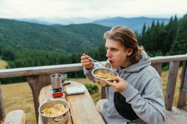 Erkek turist, dağlardaki ahşap bir evin terasında bir masada oturur ve güzel bir dağ manzarasının arka planında yemek yer..