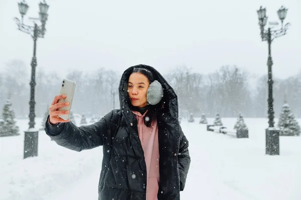 穿着外套 穿着温暖衣服的女人站在雪地的街道上 拿着智能手机自拍 女士在雪地里用智能手机相机拍照 — 图库照片