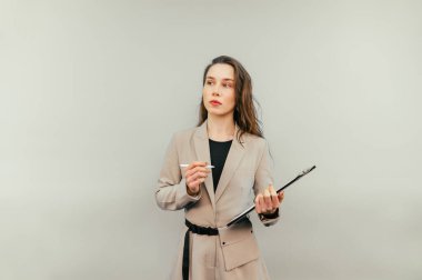 Attractive woman in a suit with a tablet and a pen in her hands stands on a beige background and looks away with a serious face.