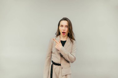 Bej ceketli şaşırmış kadın bej bir arka planda duruyor, parmağını yana doğrultuyor ve şok olmuş bir yüzle kameraya bakıyor..