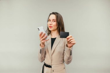 Attractive woman in a suit stands on a beige background with a smartphone and a bank card in her hands and uses the Internet, shopping online.