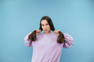 Attractive brunette woman in casual clothes holds her hands by the ponytails with her hair and poses for the camera on a blue background.