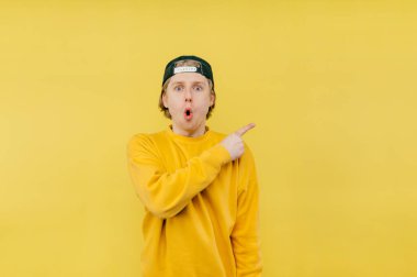 Şok olmuş, sarı kazaklı ve şapkalı adam parmağını yana doğru gösteriyor ve şaşırmış bir yüzle kameraya bakıyor..