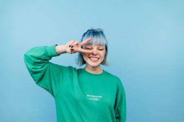 Attractive hipster girl in casual clothes stands on a blue background and shows a gesture of peace winking.