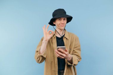 Panama ve tişörtlü komik bir genç elinde akıllı bir telefonla mavi bir arka planda duruyor ve bir jest gösteriyor tamam, gülümsüyor ve kameraya bakıyor..