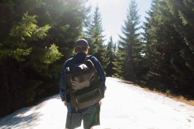 Sırtında sırt çantasıyla şık giyinmiş bir adam. Karların içinde, arka planda kozalaklı ağaçların üzerinde duruyor..