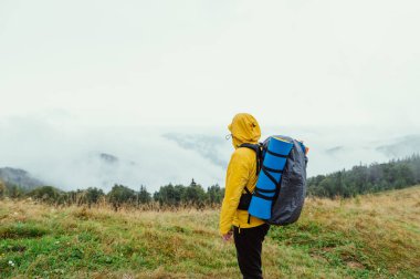 Hiker in a yellow jacket stands in the mountains on a rainy foggy day during a hike, looks at the beautiful views of nature and looks ahead