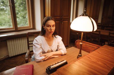 Halk kütüphanesindeki bir masada elinde kitapla oturan ve ciddi bir yüzle kameraya bakan çekici bir kadın..